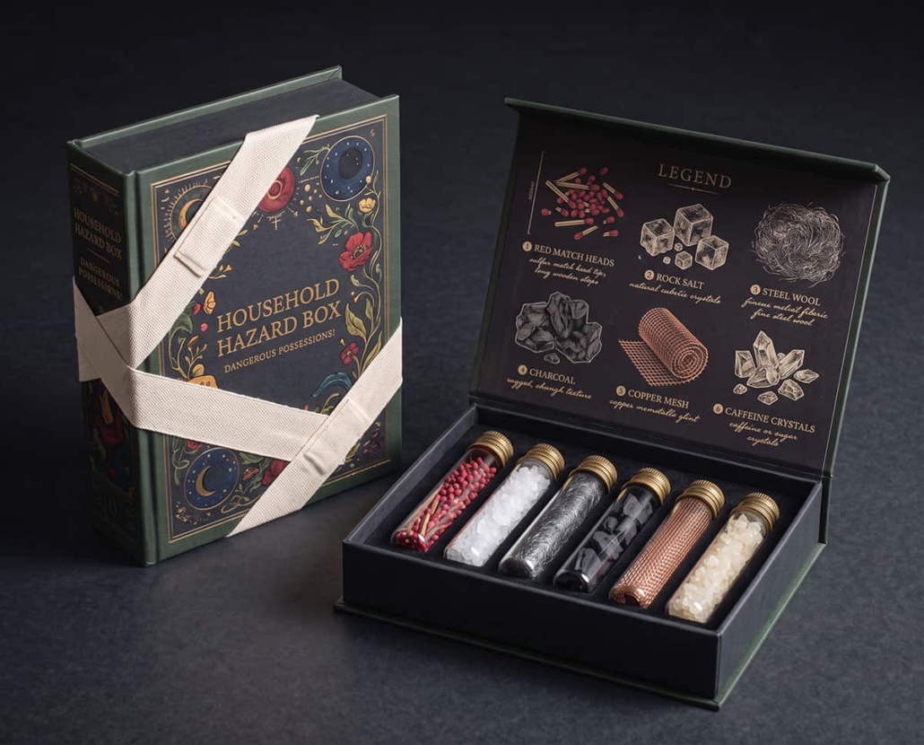 the-household-hazard-box Luxe boekvormige verpakking met decoratieve illustraties en een grote stoffen pleister rondom de doos, vormgegeven als een stijlvolle veiligheidskit voor alledaagse huishoudelijke materialen.